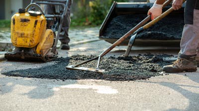Low angle view of two workers arranging fresh asphalt mix with rakes and shovel to patch a bump in the road with machinery by their side.